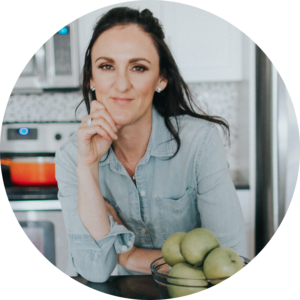 A woman in a denim shirt smiles at the camera, resting her chin on her hand, with a bowl of green apples in front of her in a kitchen.