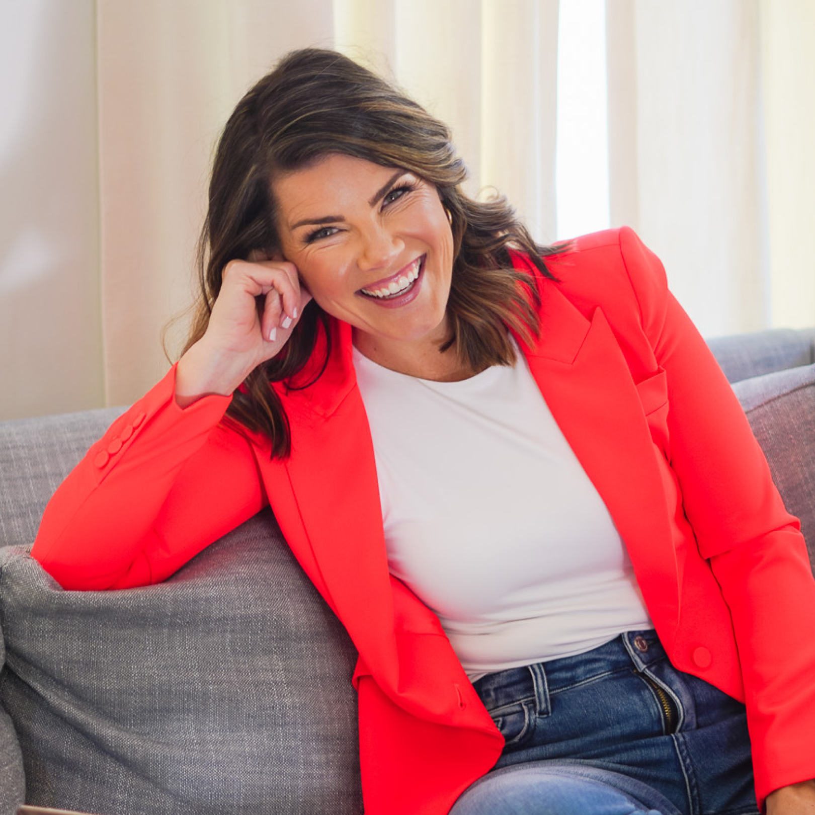 Woman in a red blazer and jeans smiling while seated on a gray couch.