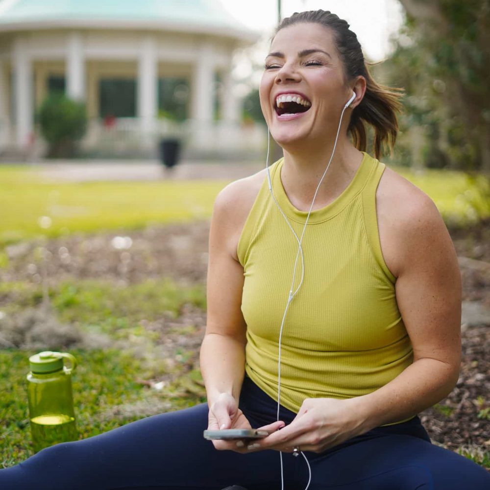 Woman laughing while using smartphone with earphones in a park.