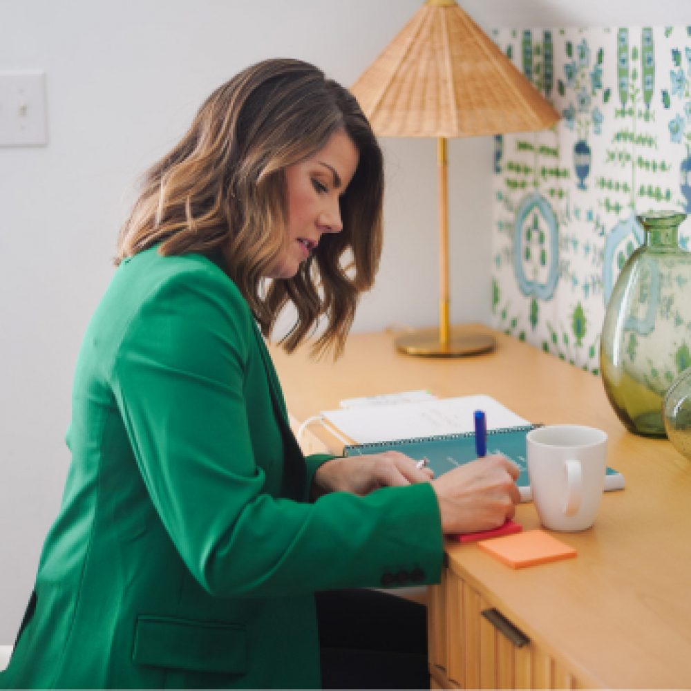 A woman in a green blazer sits at a desk, writing in a notebook next to a white cup and under a table lamp, with a patterned green curtain in the background.