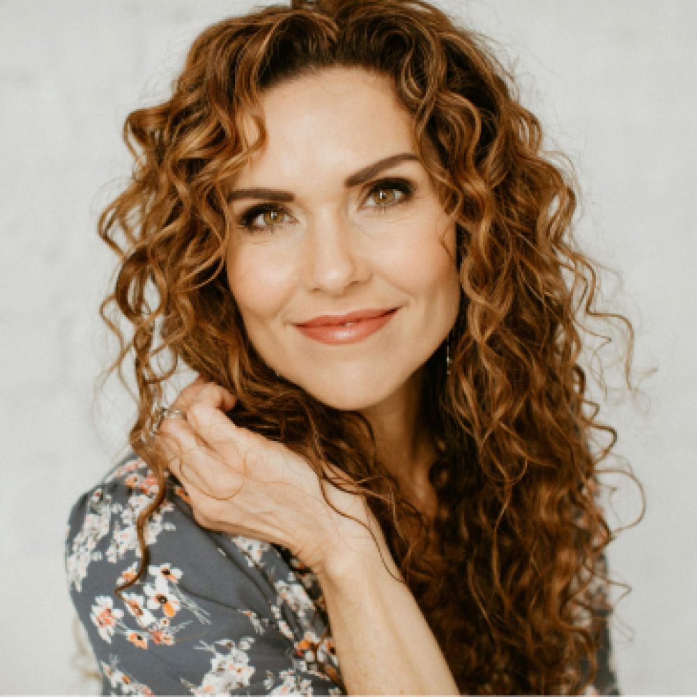 A woman with curly hair and a floral top smiling gently at the camera against a light background.