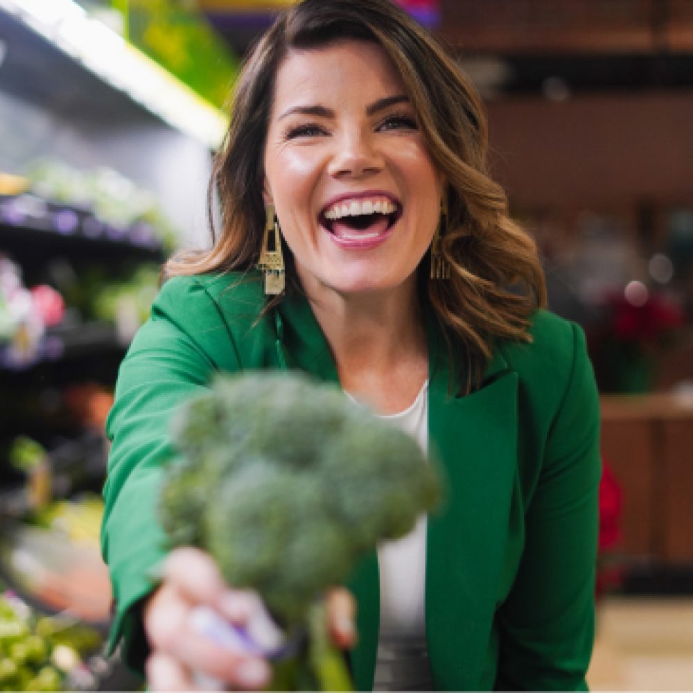 A smiling woman in a green jacket offering broccoli to the camera in a grocery store.