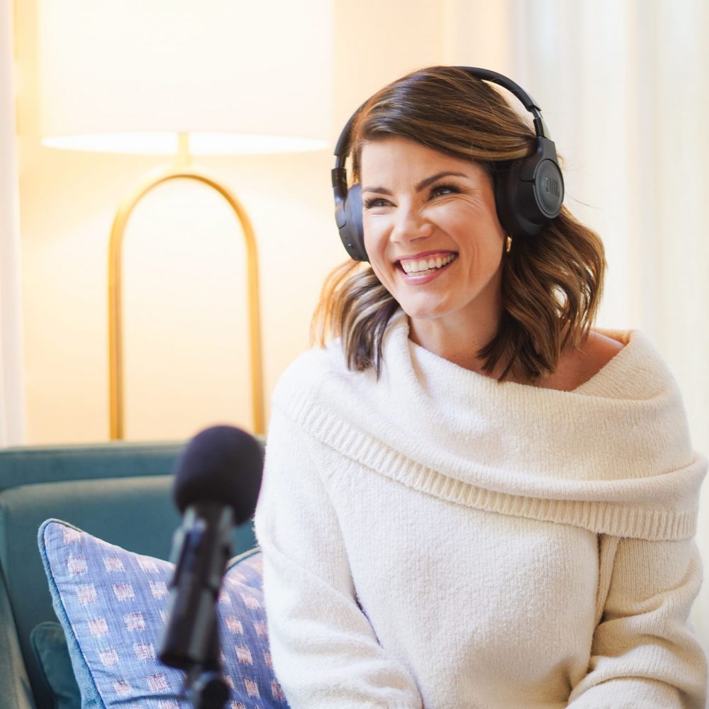 A smiling woman wearing headphones and an off-white sweater, sitting in a cozy room with a microphone and a lamp in the background.