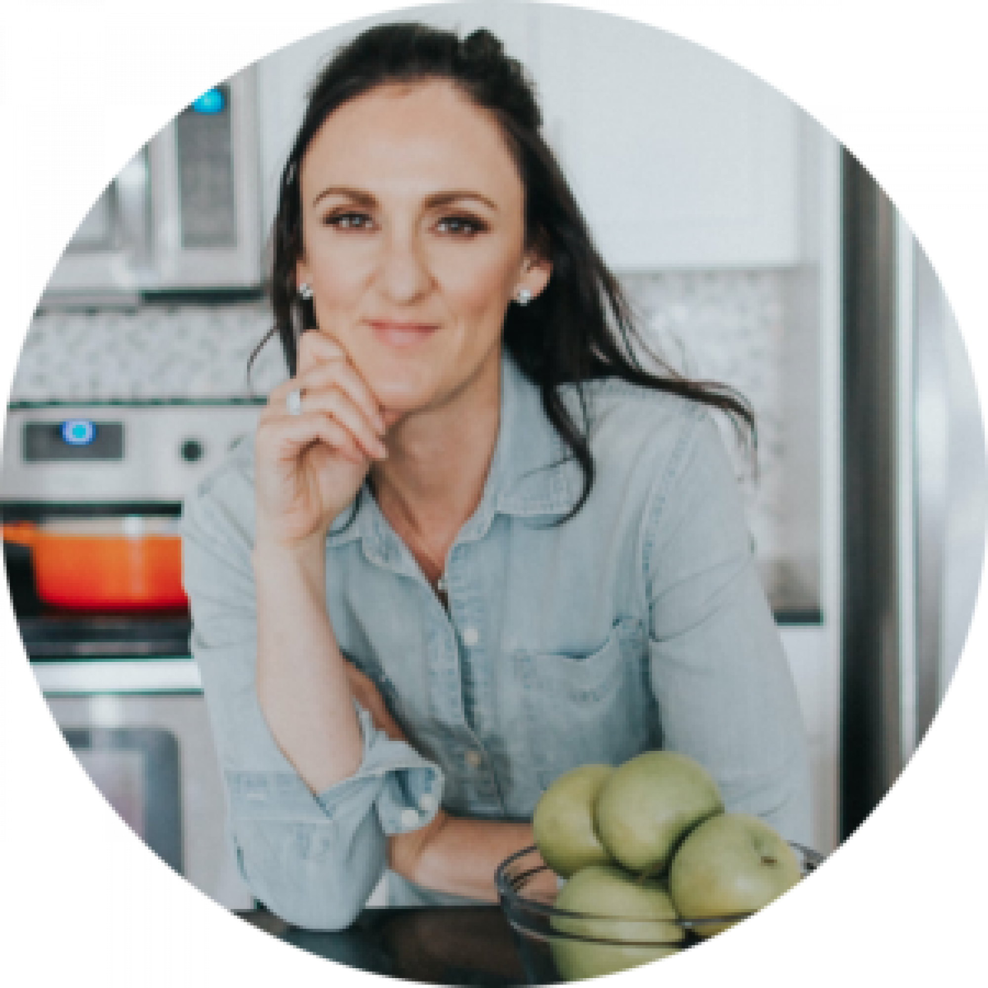 A woman in a denim shirt smiles at the camera, resting her chin on her hand, with a bowl of green apples in front of her in a kitchen.