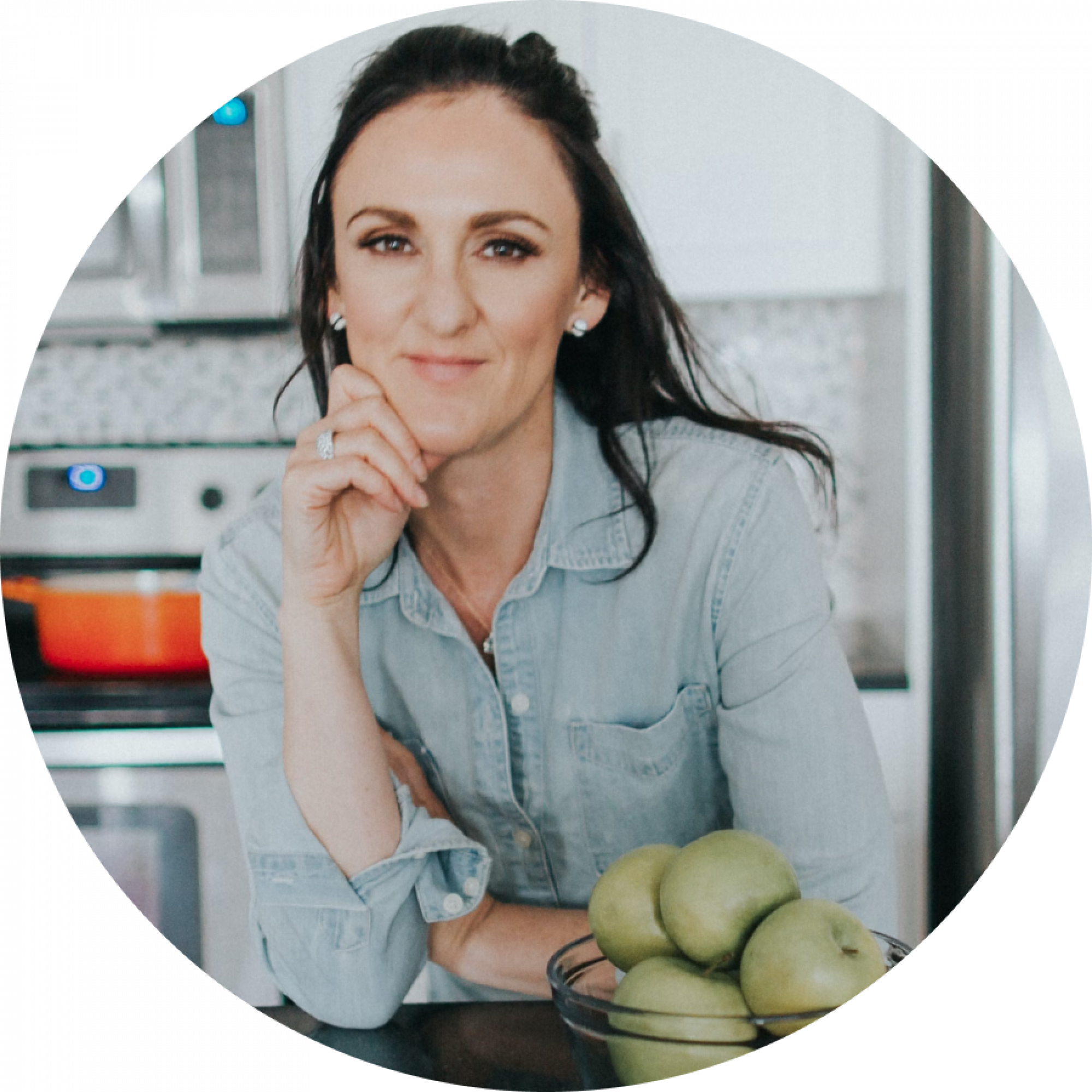 A woman in a denim shirt smiles at the camera, resting her chin on her hand, with a bowl of green apples in front of her in a kitchen.
