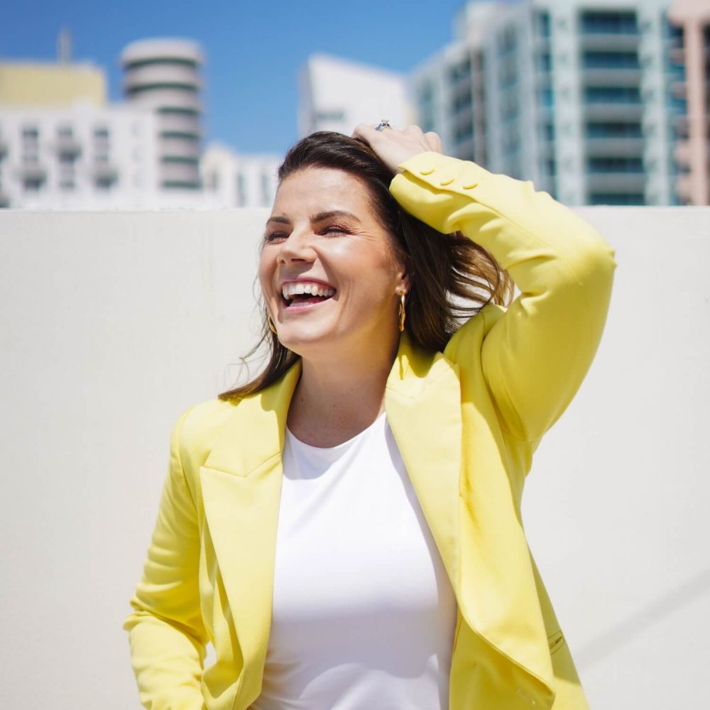 A woman in a yellow blazer and white top, smiling brightly, stands against a backdrop of skyscrapers under a clear blue sky.