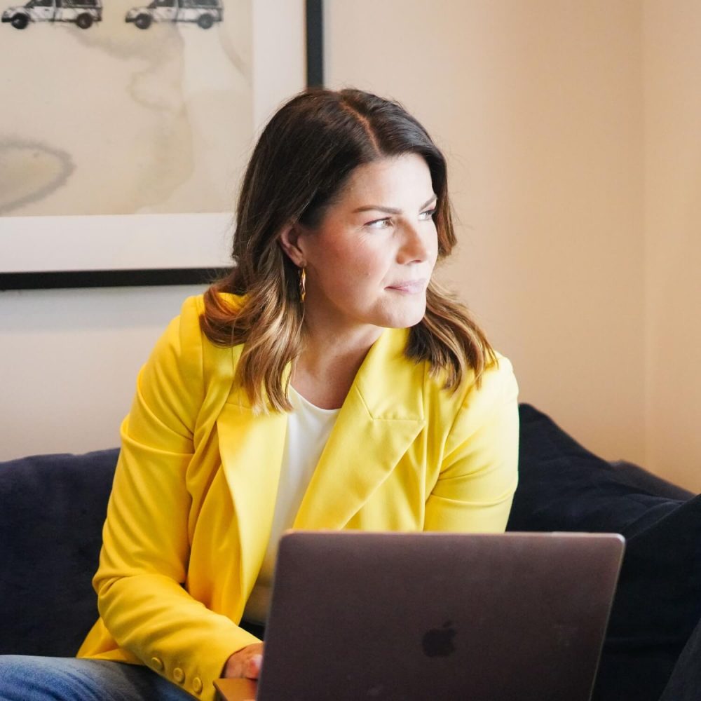 A woman in a yellow blazer sits on a couch working on a laptop, looking intently to her left with a thoughtful expression.