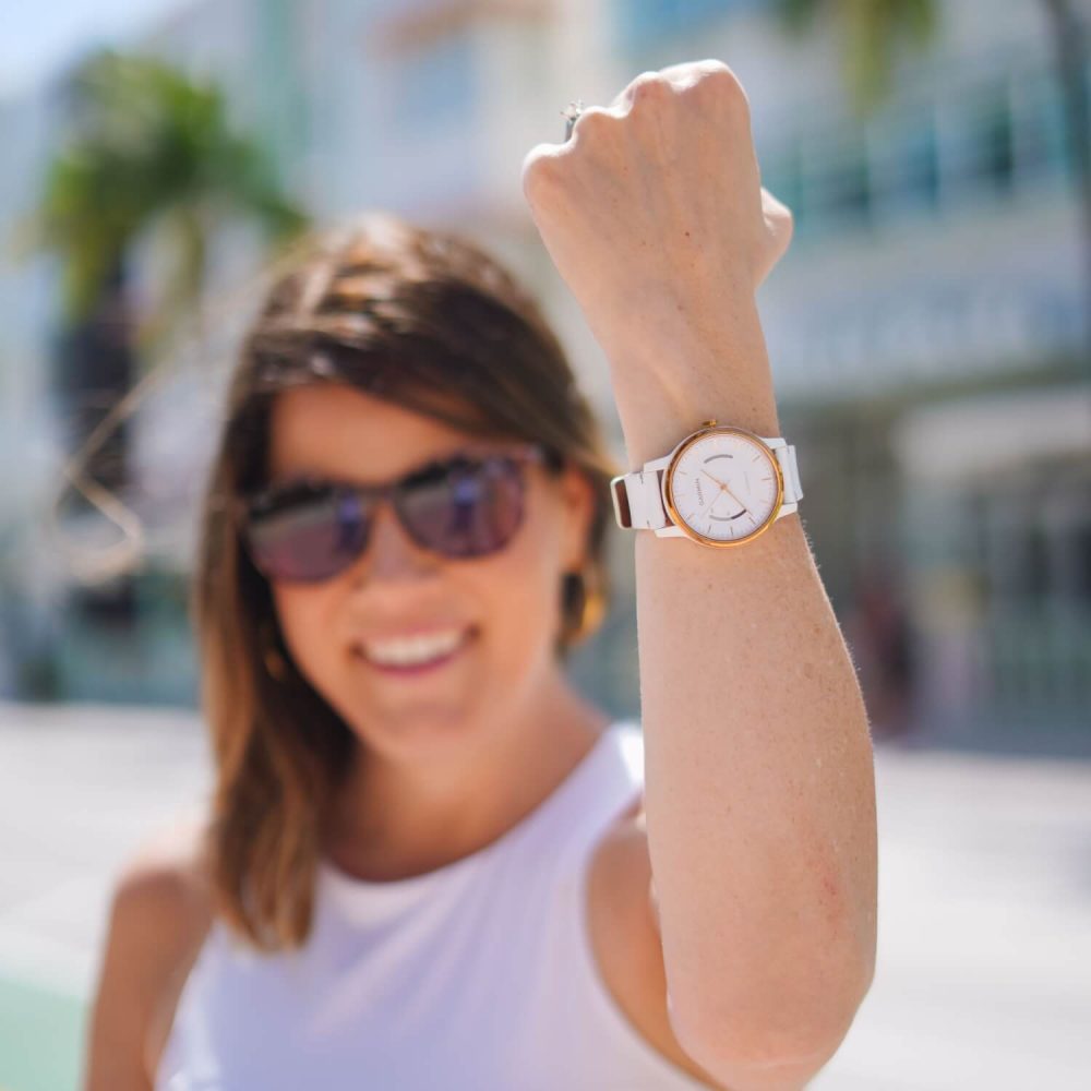 A woman in sunglasses showing off a wristwatch, smiling, with blurred urban background.